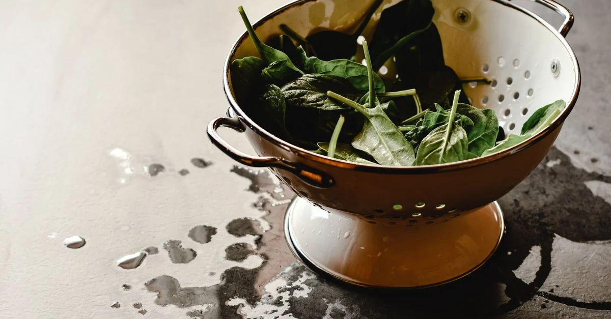 Une passoire vintage remplie de feuilles d'épinards fraîches et humides sur un plan de travail de cuisine.