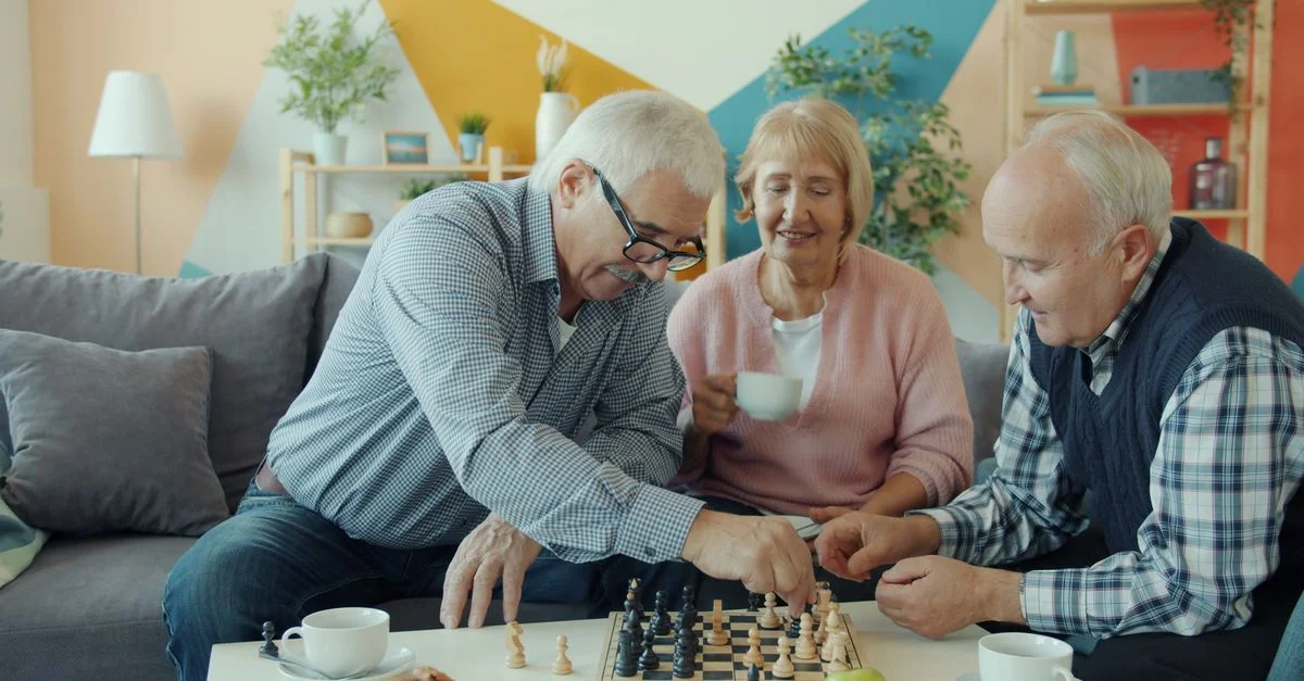 Trois personnes âgées jouent aux échecs dans un salon confortable, appréciant la compagnie les unes des autres.