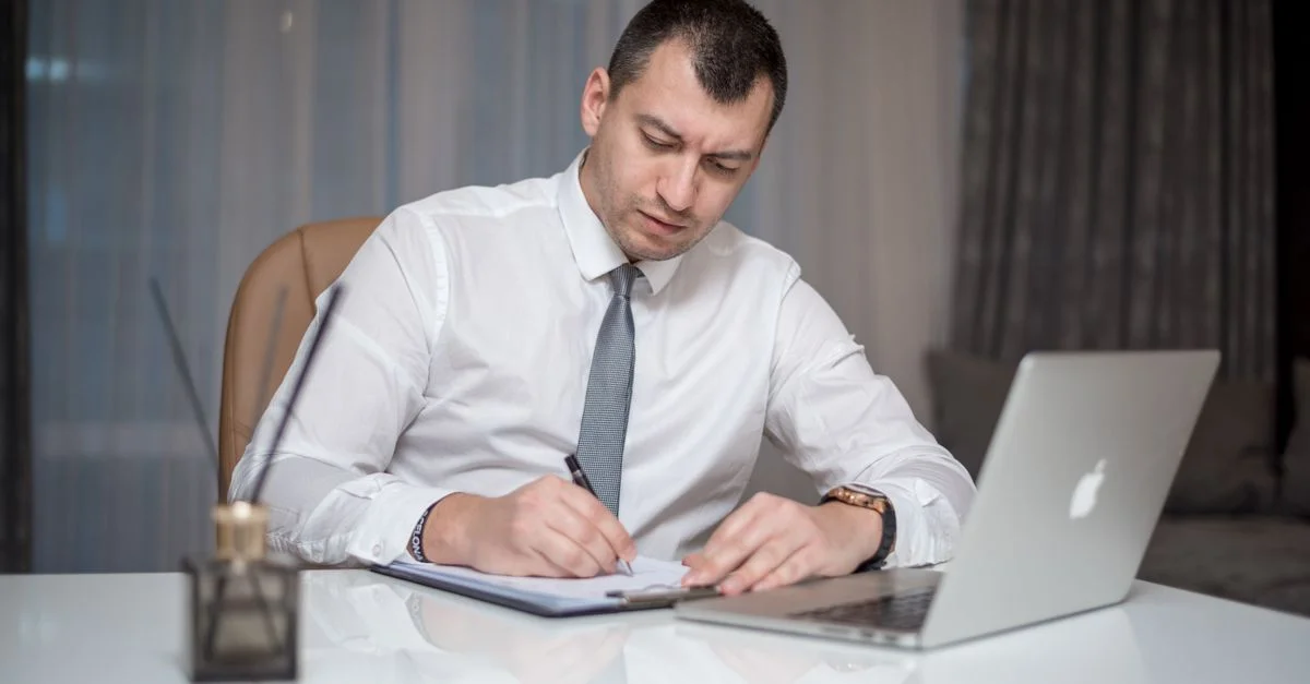 Homme d'affaires en costume prenant des notes au bureau avec un ordinateur portable, reflétant la vie moderne au bureau.